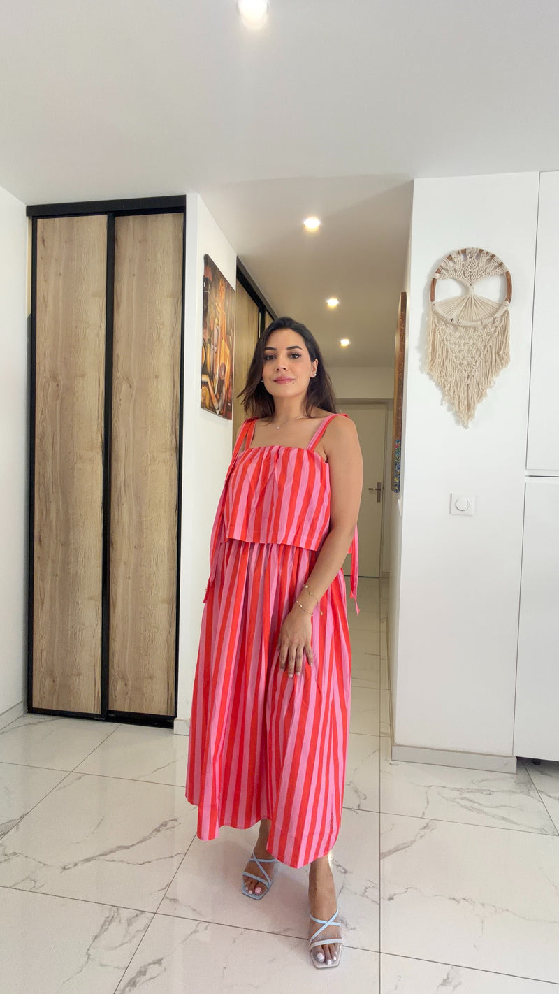 Femme portant une robe longue fluide rayée rose et corail avec bretelles nouées, accessoirisée d'un chapeau de paille et d'un sac en raphia